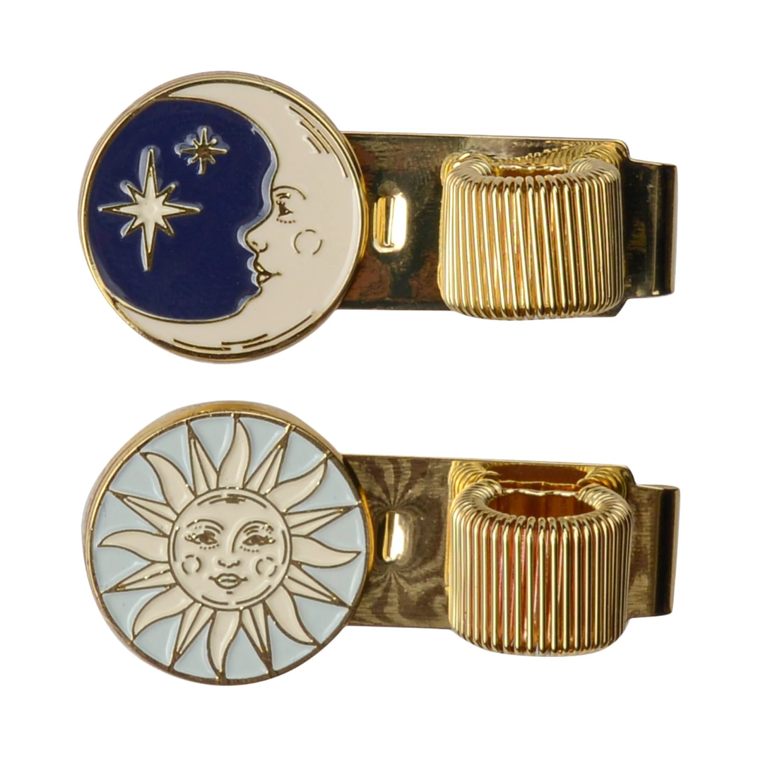 Two clips, one a circle of a moon and stars in off-white and navy blue next to a gold loop for the pen, over a circle of an off-white sun on light blue background next to its pen loop