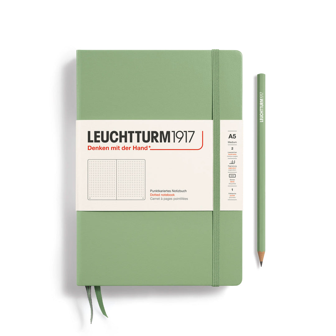 A Leuchtturm1917 A5 hardcover dotted page notebook in sage green with a white label on the front, shown closed with an elastic closure and ribbon bookmark. A matching sage green pencil is placed vertically to the right.