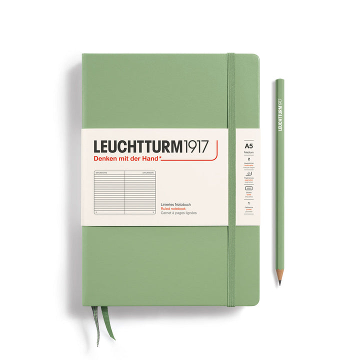 A Leuchtturm1917 A5 hardcover ruled page notebook in sage green with a white label on the front, shown closed with an elastic closure and ribbon bookmark. A matching sage green pencil is placed vertically to the right.