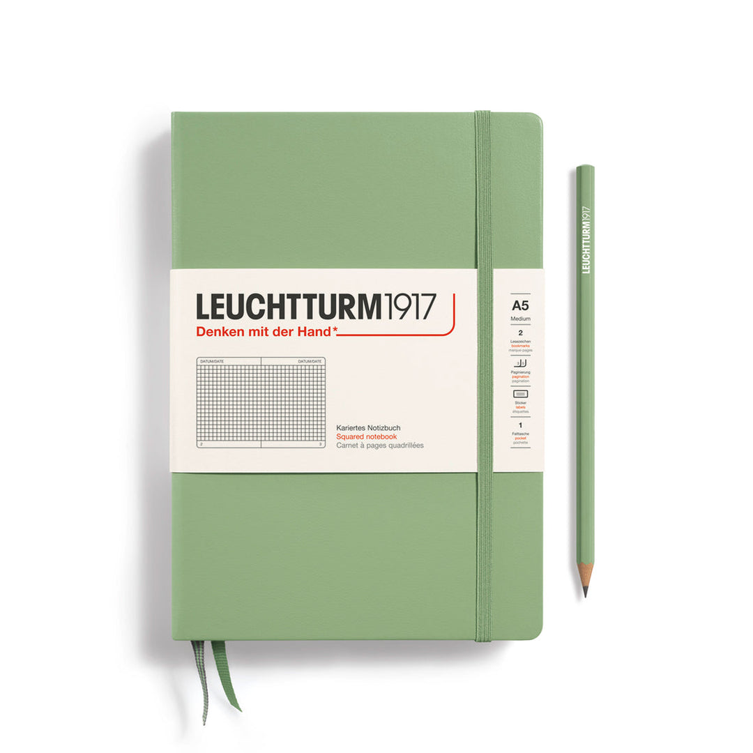 A Leuchtturm1917 A5 hardcover squared page notebook in sage green with a white label on the front, shown closed with an elastic closure and ribbon bookmark. A matching sage green pencil is placed vertically to the right.