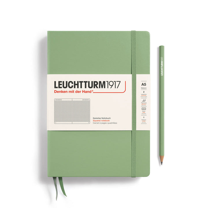 A Leuchtturm1917 A5 hardcover squared page notebook in sage green with a white label on the front, shown closed with an elastic closure and ribbon bookmark. A matching sage green pencil is placed vertically to the right.