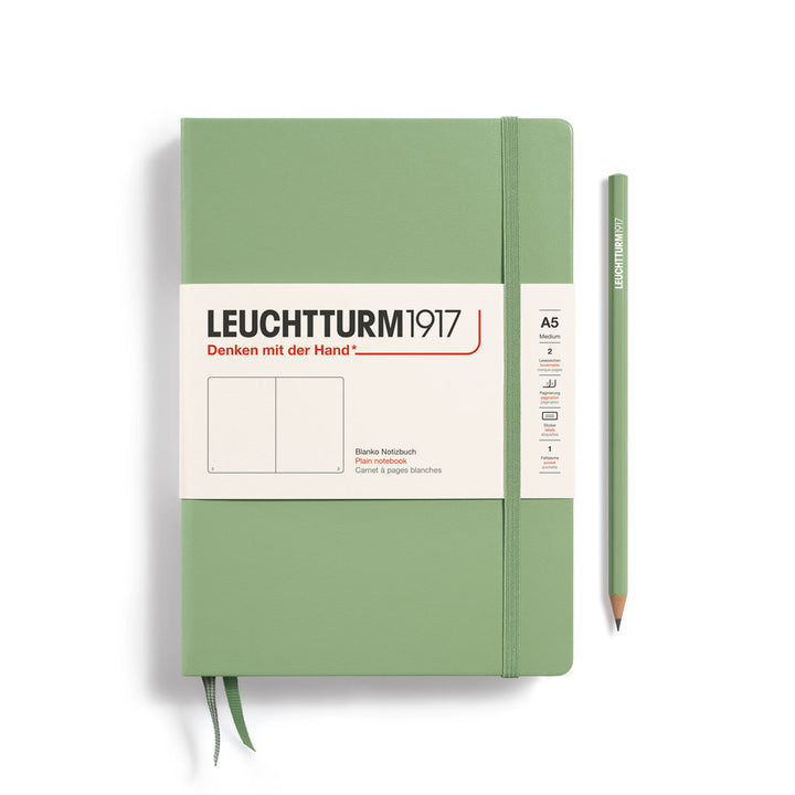 A Leuchtturm1917 A5 hardcover plain page notebook in sage green with a white label on the front, shown closed with an elastic closure and ribbon bookmark. A matching sage green pencil is placed vertically to the right.