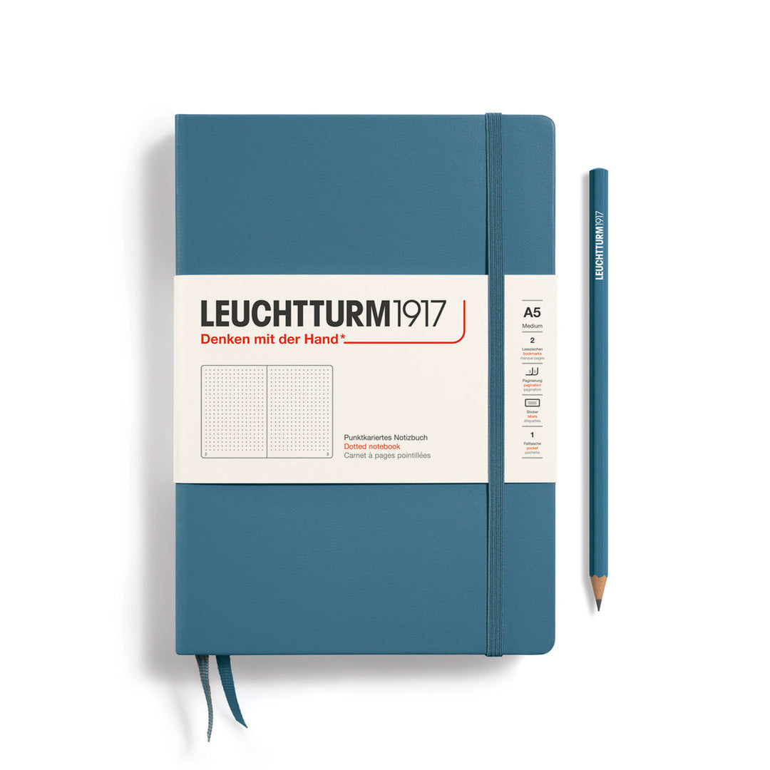 A Leuchtturm1917 A5 hardcover dotted page notebook in stone blue with a white label on the front, shown closed with an elastic closure and ribbon bookmark. A matching stone blue pencil is placed vertically to the right.