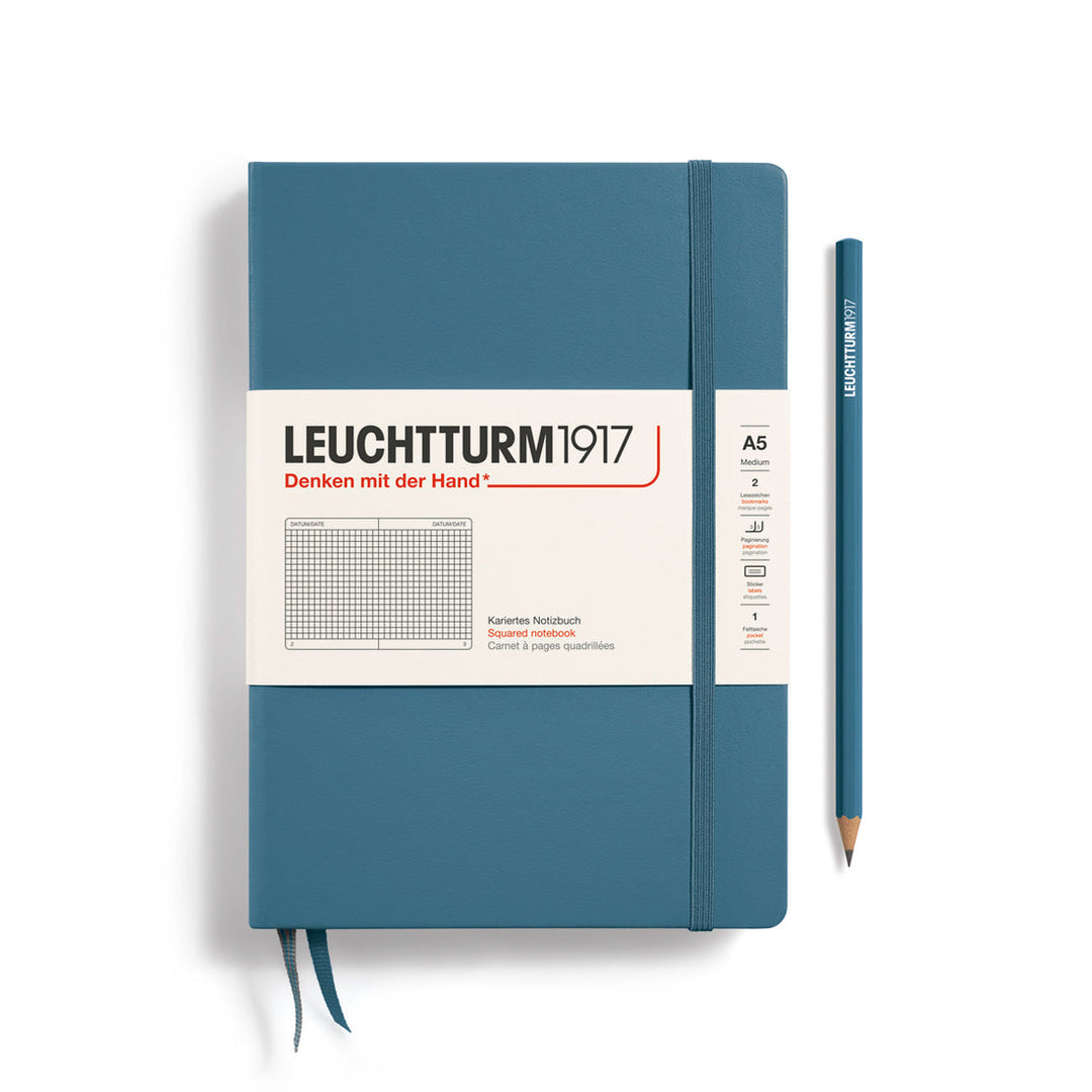 A Leuchtturm1917 A5 hardcover squared page notebook in stone blue with a white label on the front, shown closed with an elastic closure and ribbon bookmark. A matching stone blue pencil is placed vertically to the right.