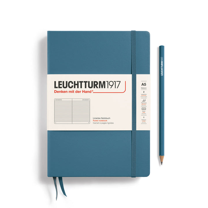 A Leuchtturm1917 A5 hardcover ruled page notebook in stone blue with a white label on the front, shown closed with an elastic closure and ribbon bookmark. A matching stone blue pencil is placed vertically to the right.