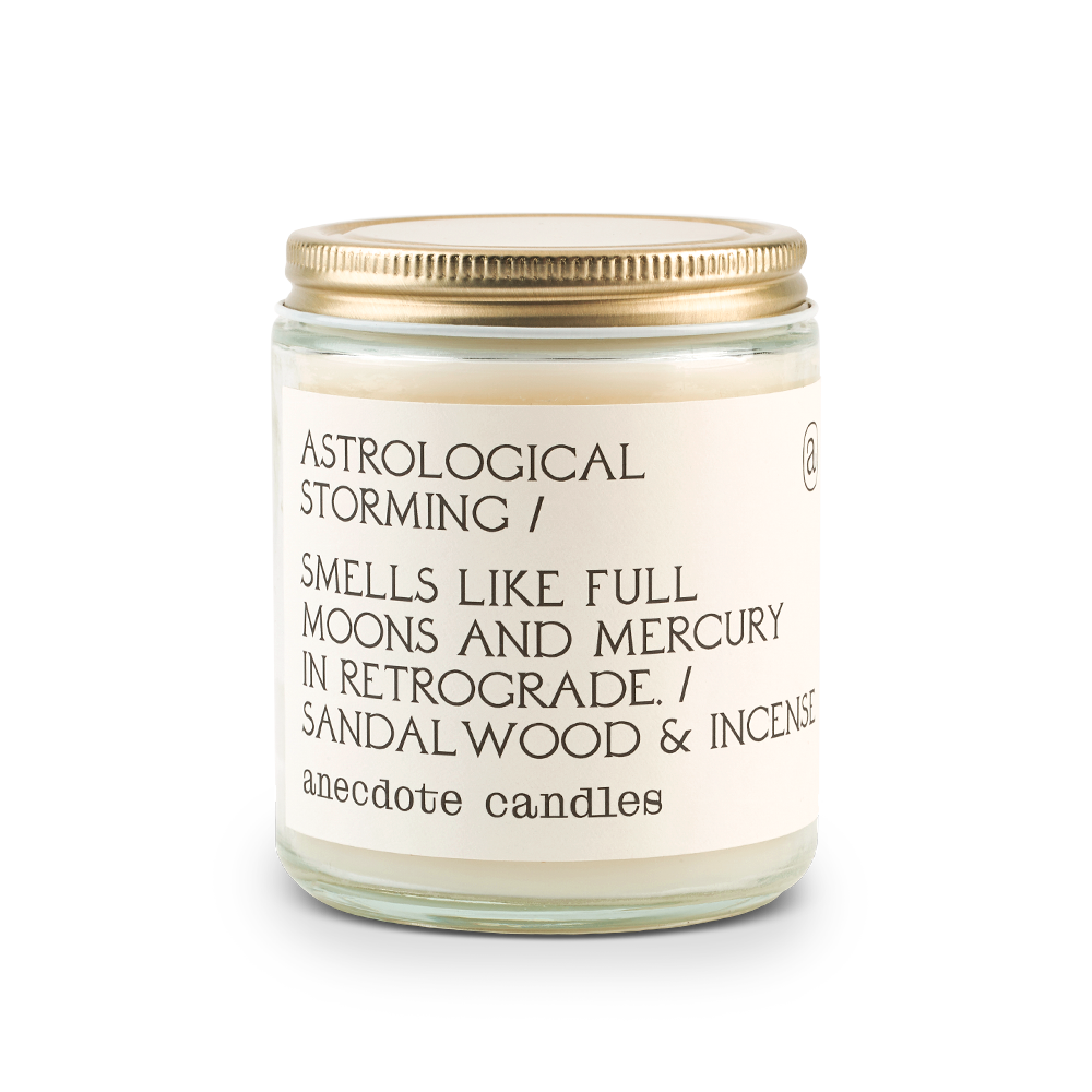 Coconut-soy wax candle in clear glass jar with lid. Label  is Astrological Storming/ Smells Like Full Moons and Mercury in Retrograde. / Sandalwood ^ Incense