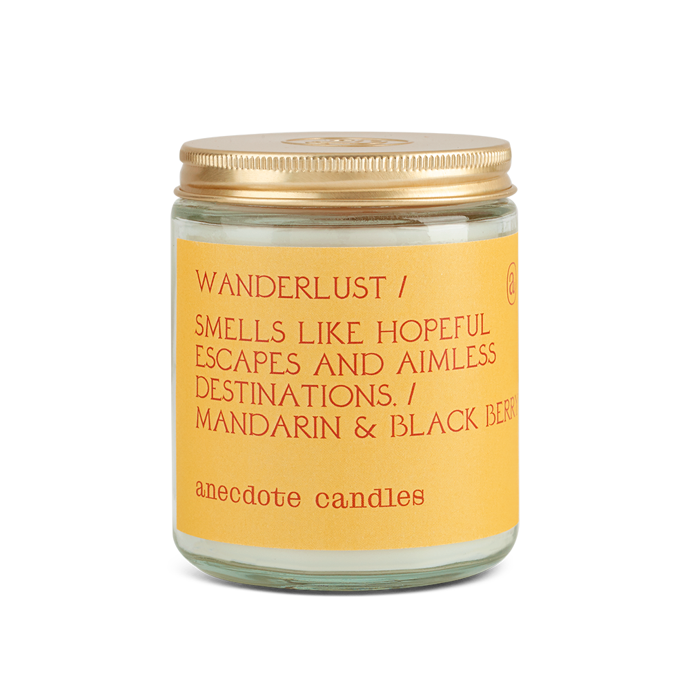 Coconut-soy wax scented candle in a clear glass jar with lid.  Yellow label with orange text is Wanderlust / Smells like Hopeful Escapes and Aimless Destinations / Mandarin & Blackberry