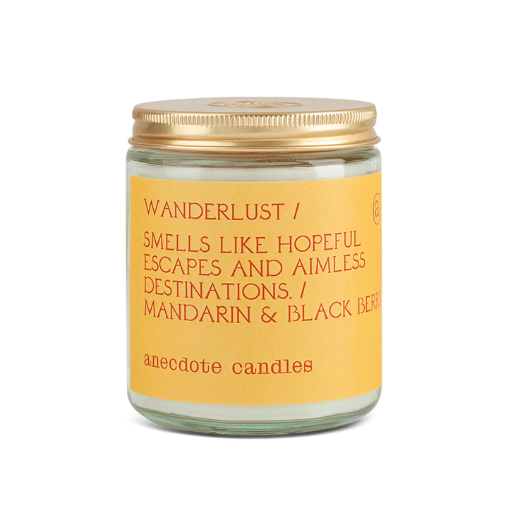 Coconut-soy wax scented candle in a clear glass jar with lid.  Yellow label with orange text is Wanderlust / Smells like Hopeful Escapes and Aimless Destinations / Mandarin & Blackberry